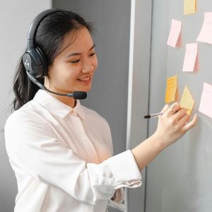 Smiling call center agent wearing headset, writing notes in office.
